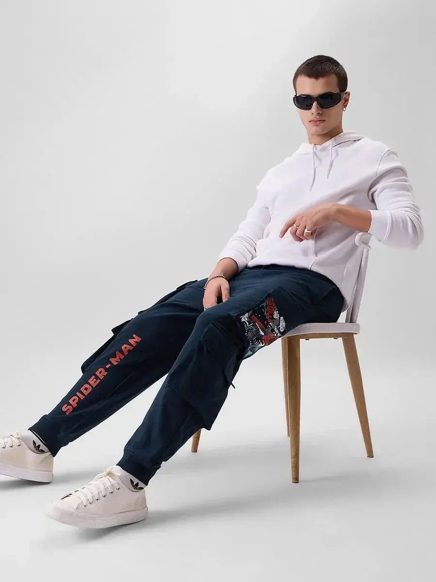 Man in white hoodie and sunglasses wearing navy baggy fit cargo joggers with Spider-Man print, sitting on chair