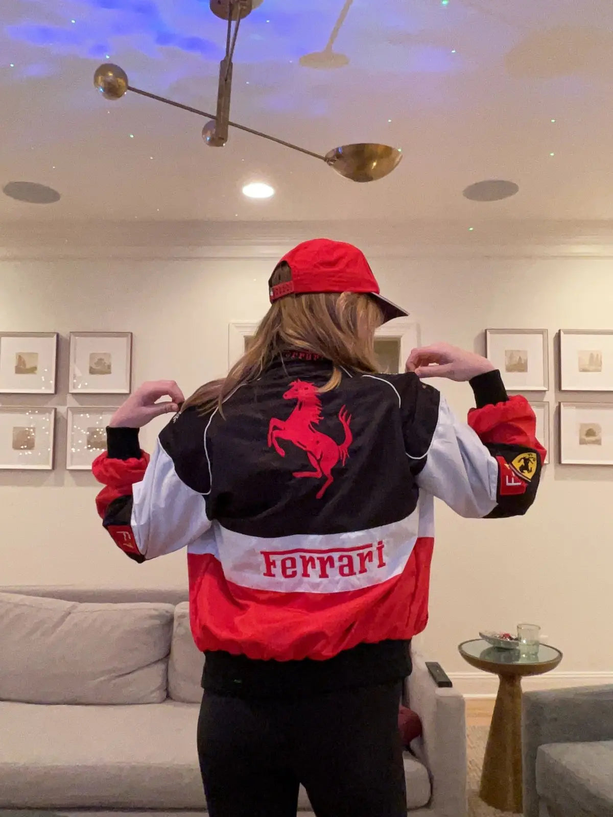 Person wearing a red and black Ferrari racing jacket and cap in a modern living room.