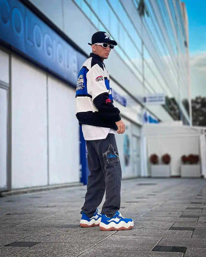 Man in streetwear blue and white varsity jacket, dark pants, chunky blue sneakers, posing outdoors