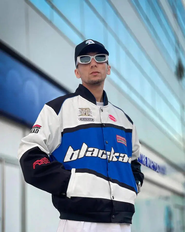 Man wearing a blue and white streetwear jacket with bold text, sporty patches, and sunglasses