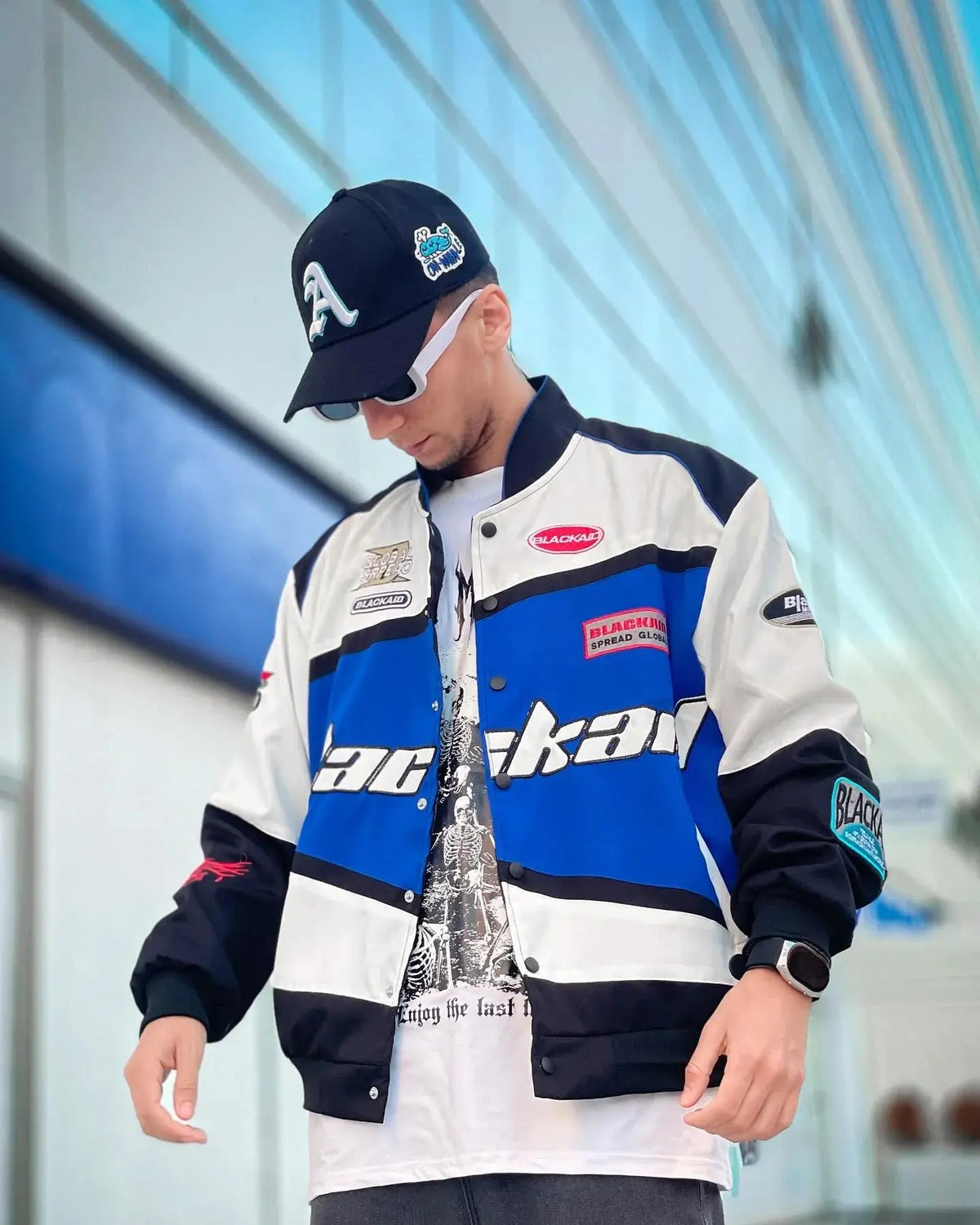 Man in blue and white streetwear jacket with bold patches, wearing cap and sunglasses outside