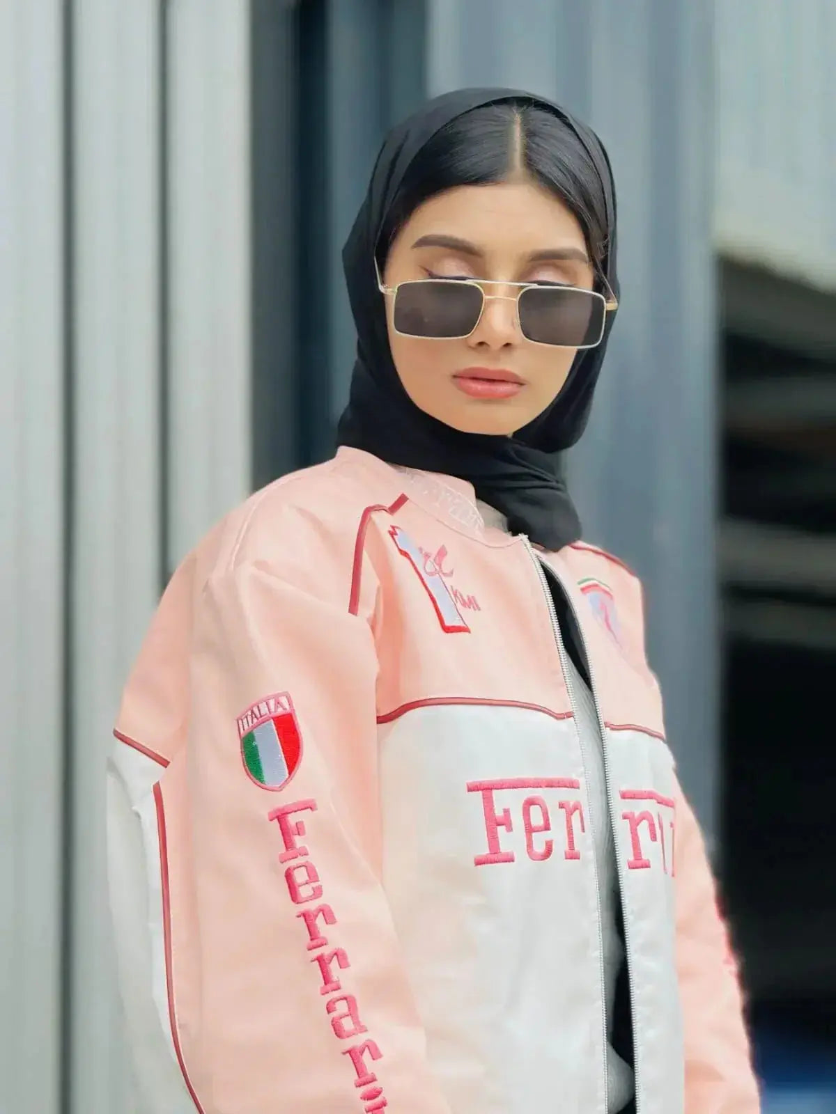 Woman wearing a pink Ferrari racing jacket, sunglasses, and a black hijab in urban setting