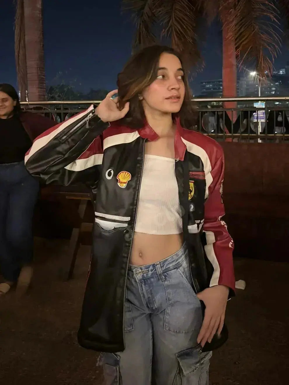 Woman in a Ferrari racing leather jacket with red, black, and white panels, wearing casual jeans, standing outdoors at night with palm trees in the background.